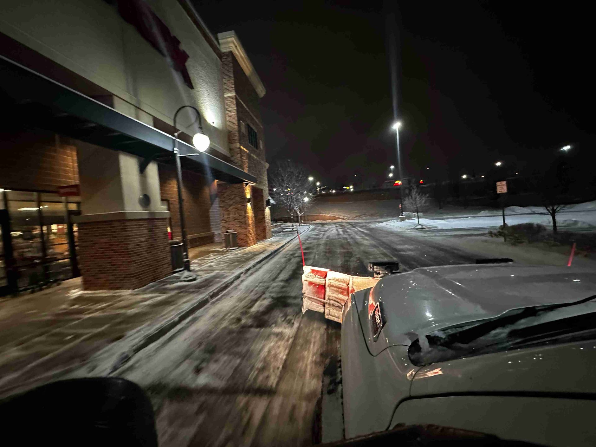 Snowy parking lot outside a building at night, seen from inside a vehicle with a plow or hood in view.