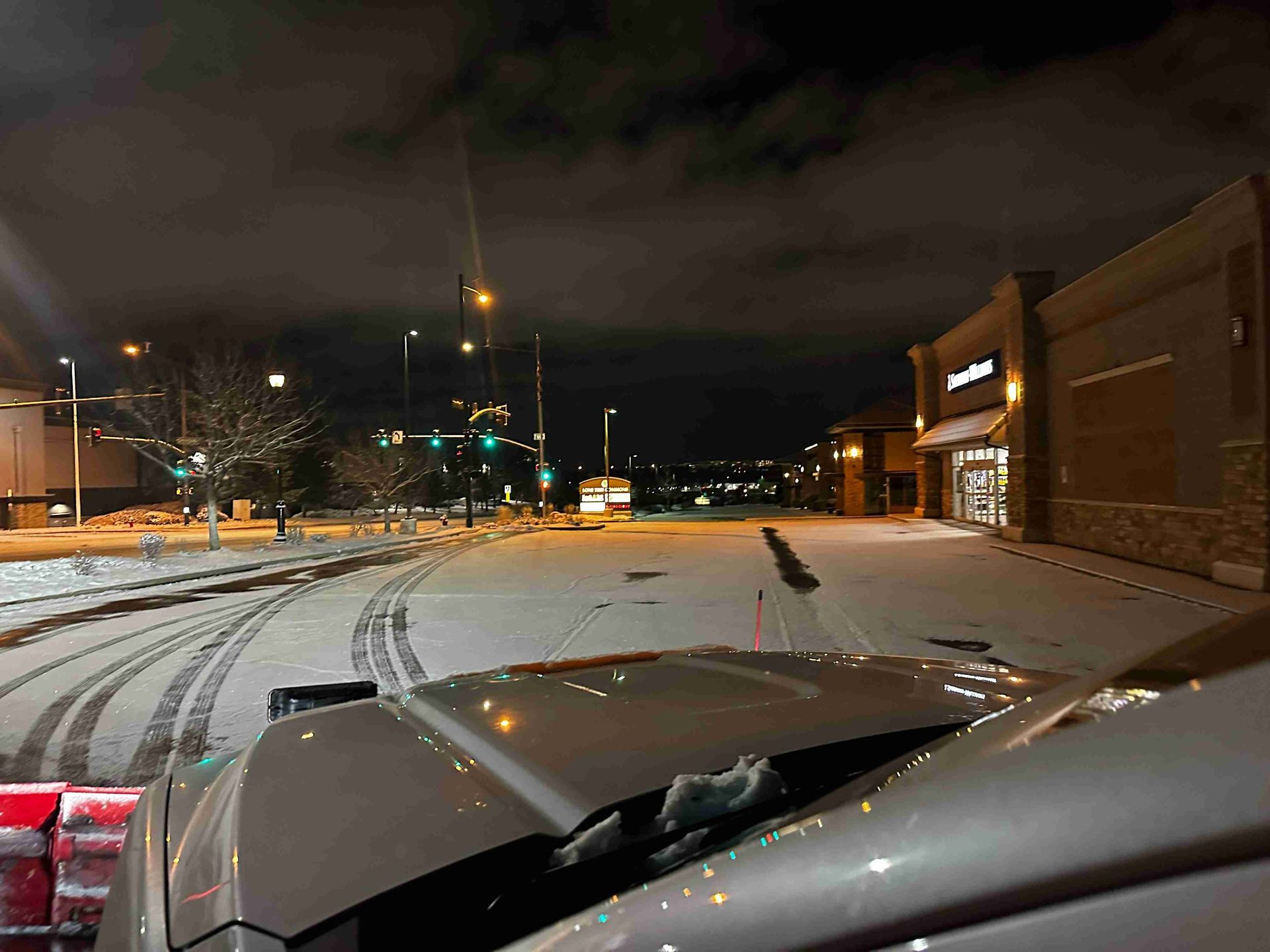 Snowy night street seen over car hood, with tire tracks, traffic lights, and lit storefronts on the right