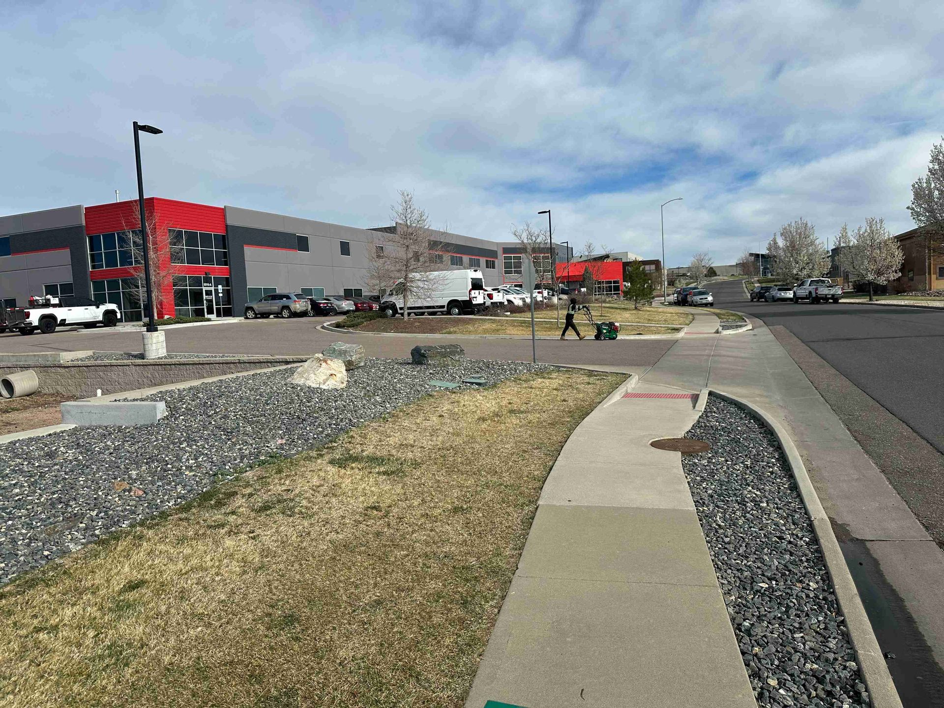 Sidewalk beside a parking lot and red commercial building on a cloudy day, with landscaped gravel and grass