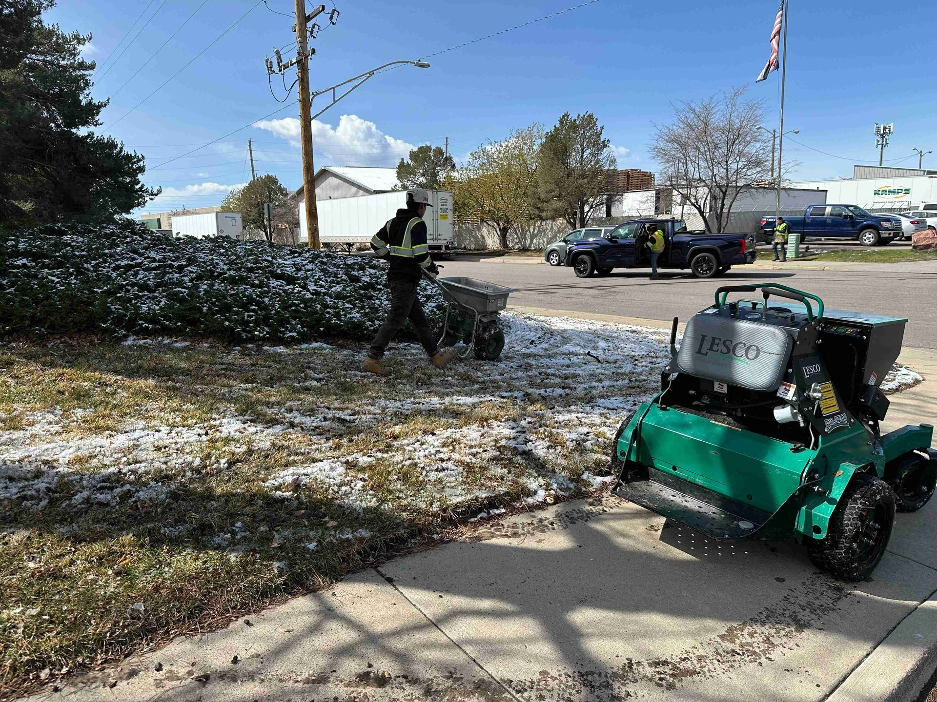 Person using a green leaf blower in a snowy parking lot with cars and a flagpole in the background