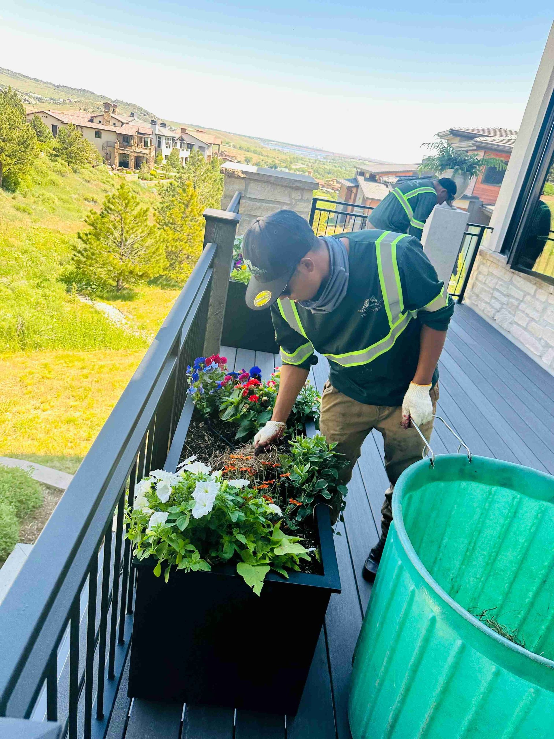 Person watering flowers on an apartment balcony beside a green bucket and railing