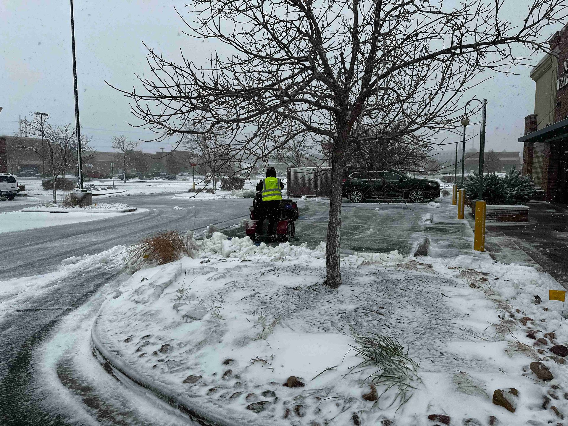 Snowy parking lot with a person on a red snow blower clearing a path near a tree and building.