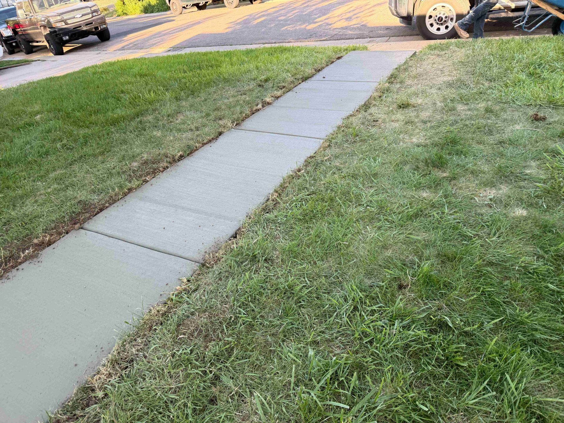 Sidewalk cutting through a grassy yard toward a street with parked cars in the background