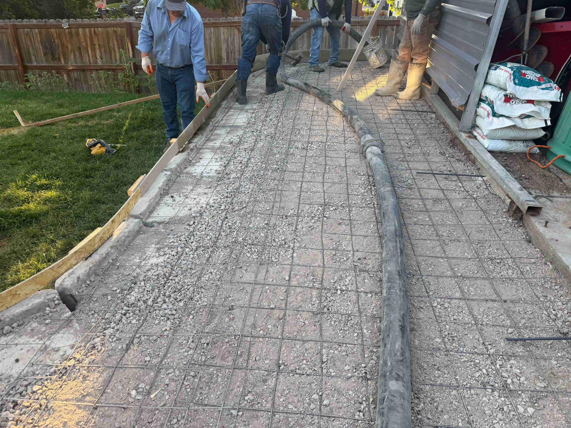 Workers pouring concrete on a curved sidewalk beside a fence and yard.