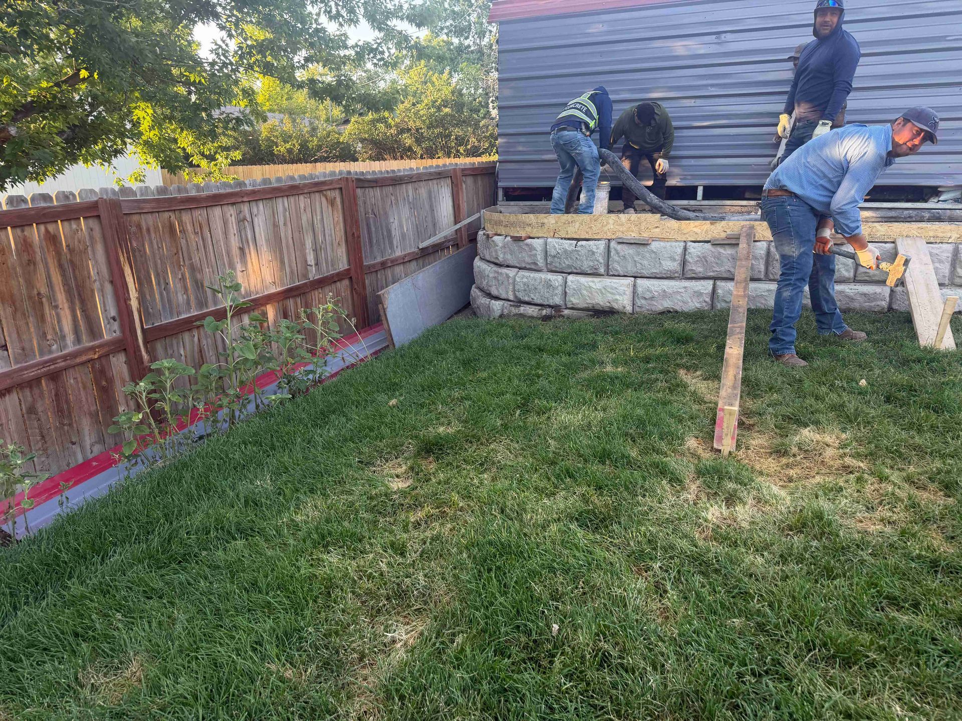 Workers building a stacked stone retaining wall beside a wooden fence in a grassy backyard