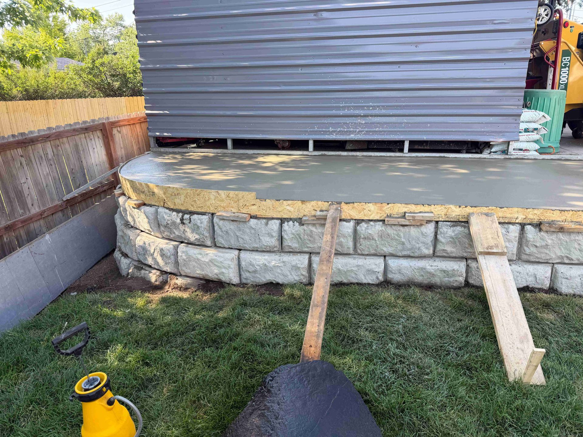 Stone retaining wall supporting a raised shed, with dirt, grass, and wooden planks in front.