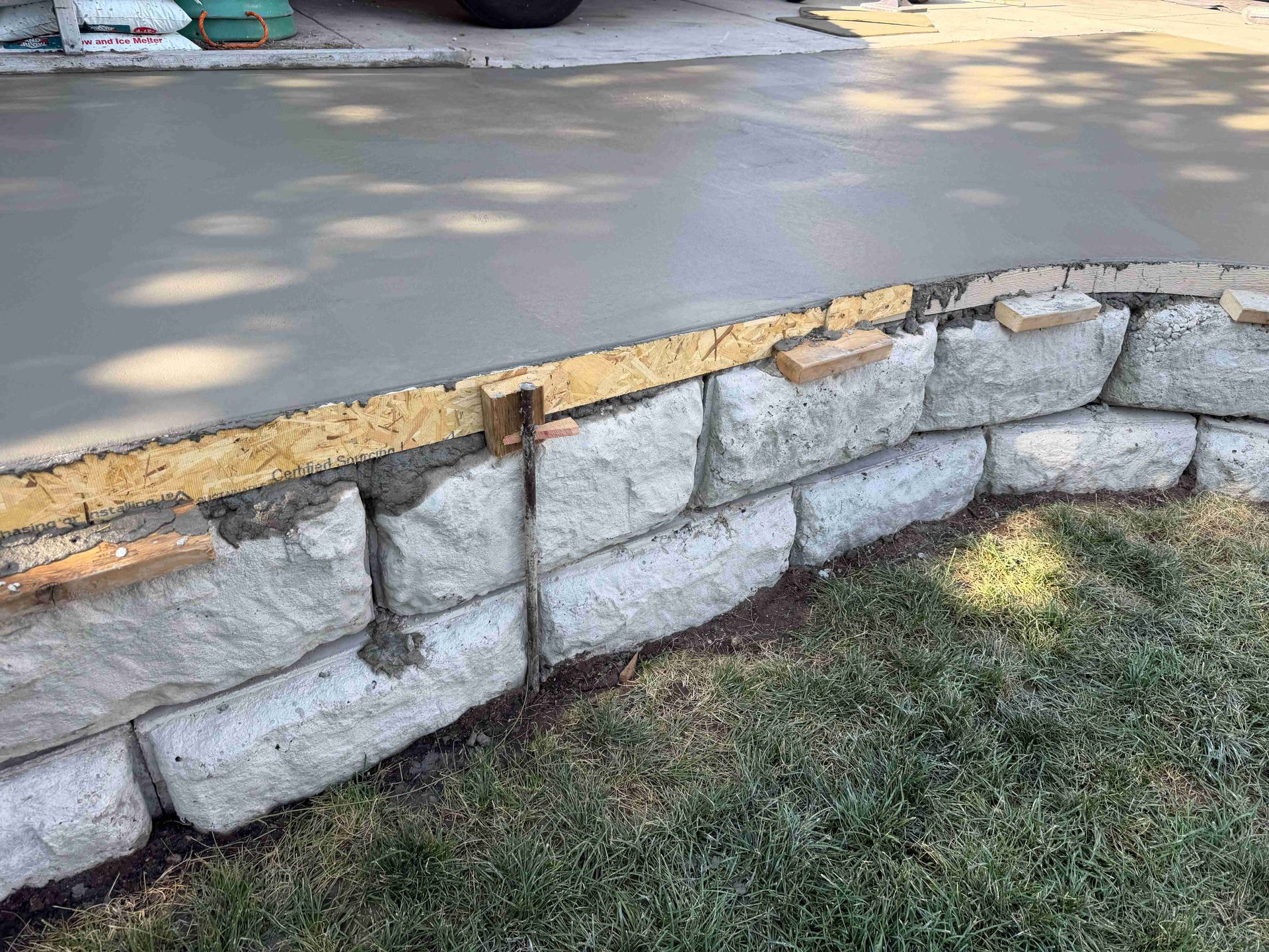 Stacked stone retaining wall bordering a paved road beside grass, with a broken wooden top edge.