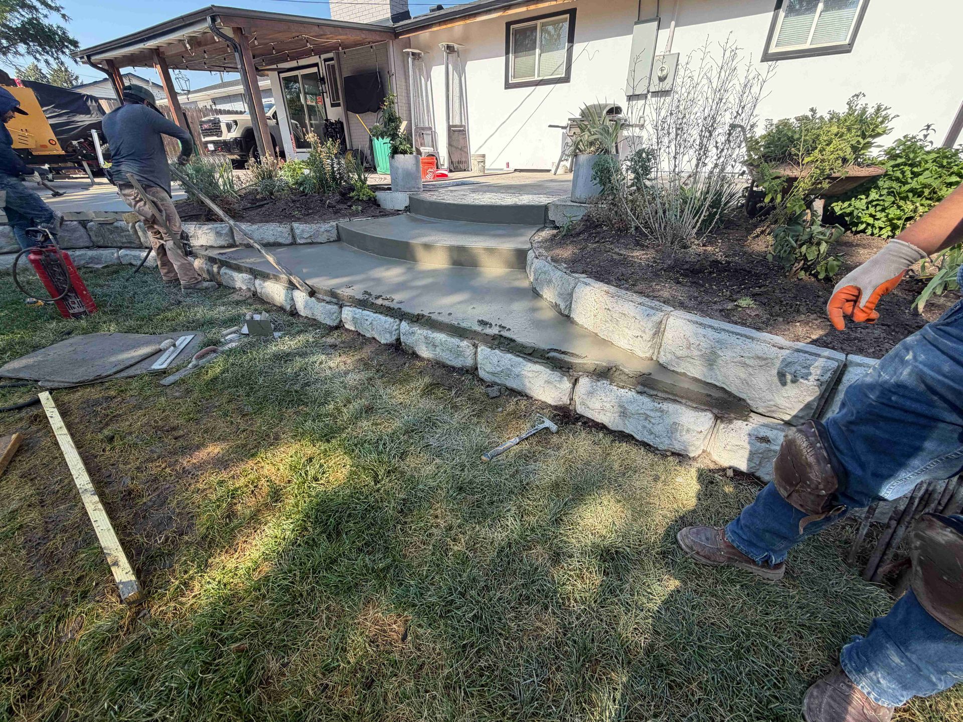 Workers building a concrete front step and retaining wall beside a house, with tools and landscaping materials nearby.
