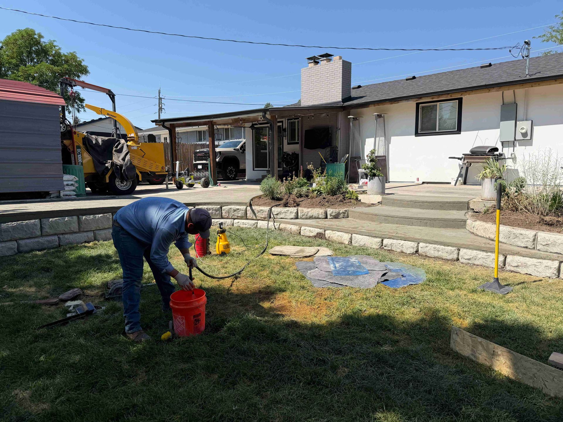 Person bending over in a backyard, filling a red bucket near a house and garden beds.