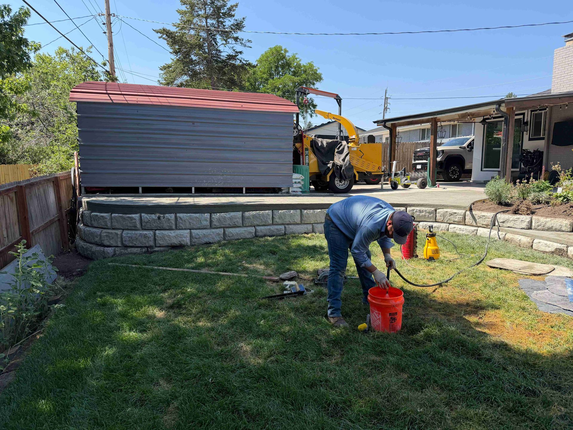 Worker kneeling on lawn with red bucket and hose near a shed and construction equipment in a backyard