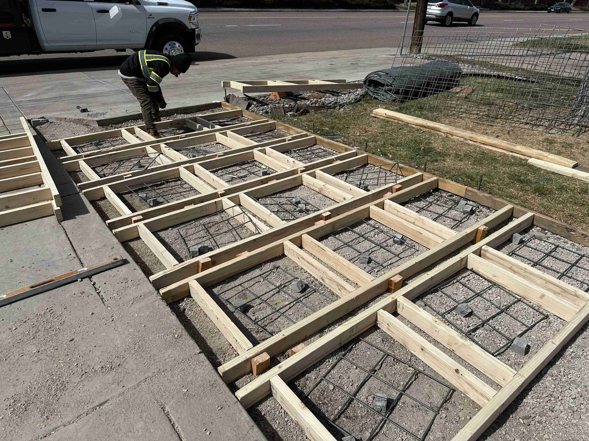 Wooden formwork laid out on a sidewalk for a concrete project, with a worker in the background.