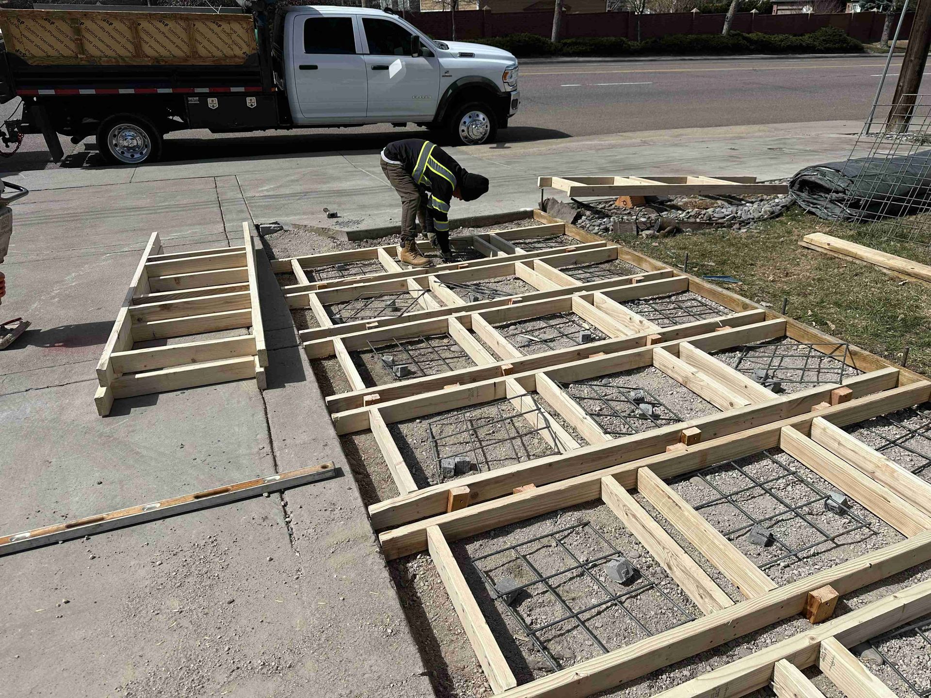 Worker assembling wooden formwork on a driveway with a power drill near a pickup truck.