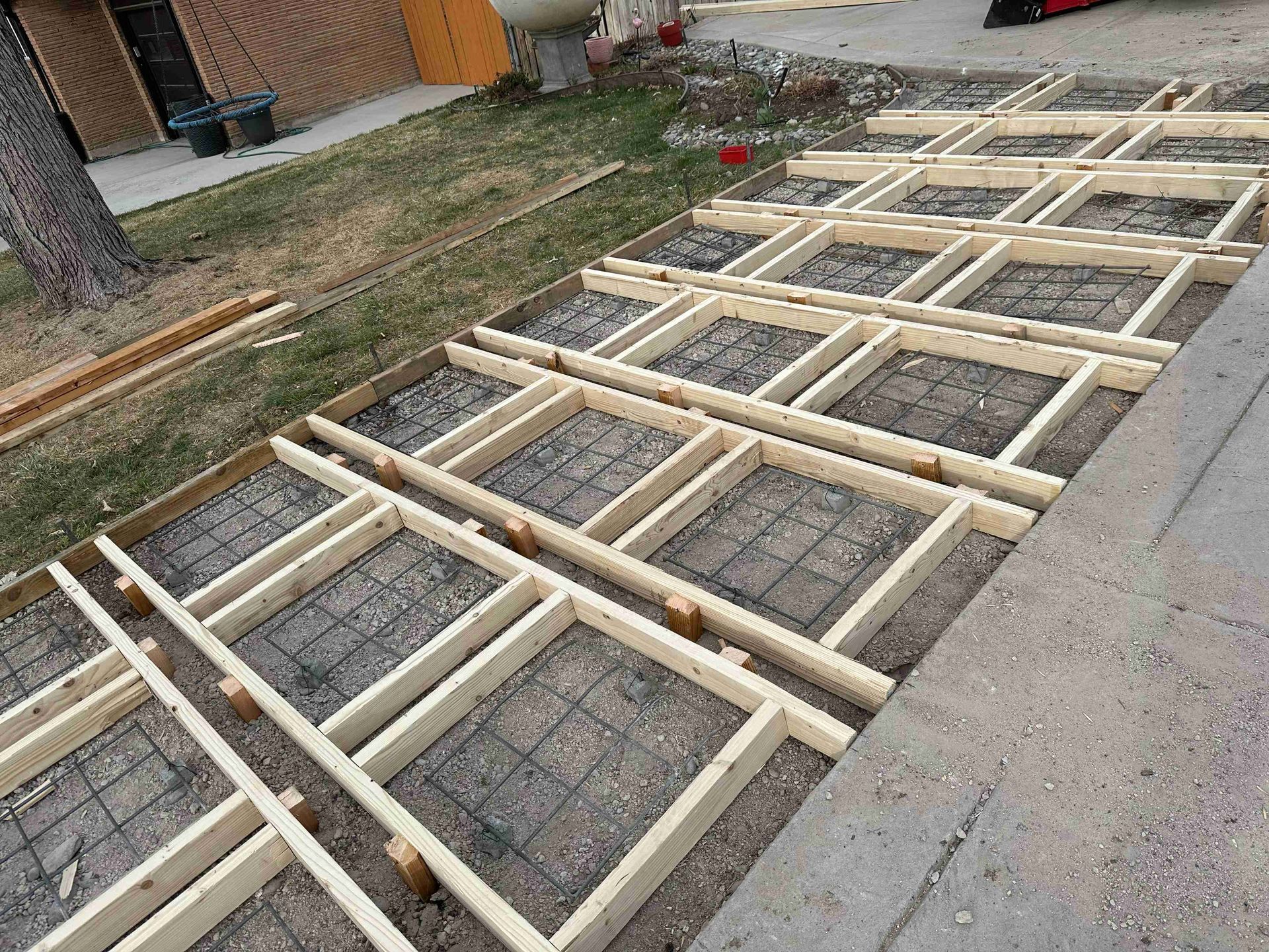 Wooden frame grid laid on a sidewalk beside a road, with concrete blocks inside each section.