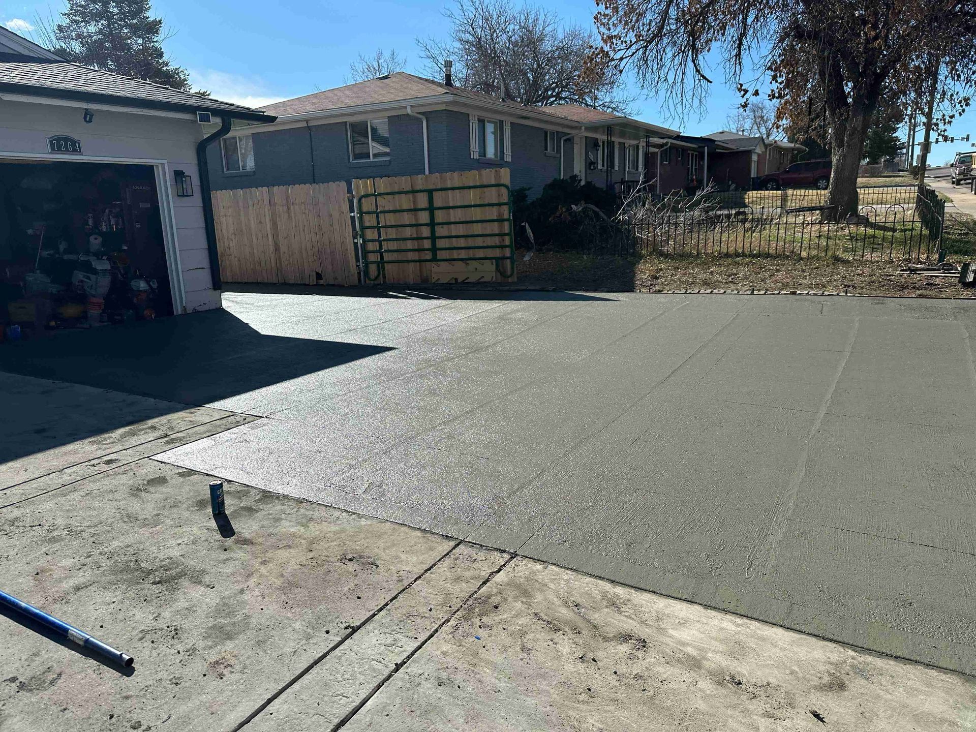 Concrete driveway leading to a single-story house with a fenced yard and trees in front.