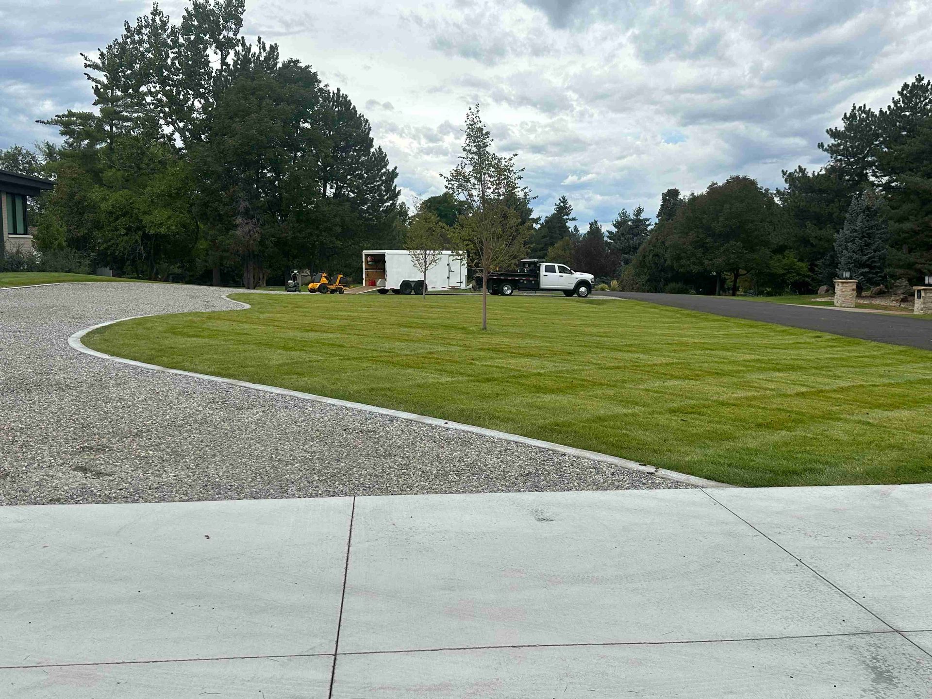 Gravel driveway curves past a grassy lawn to parked trucks and trees under a cloudy sky.