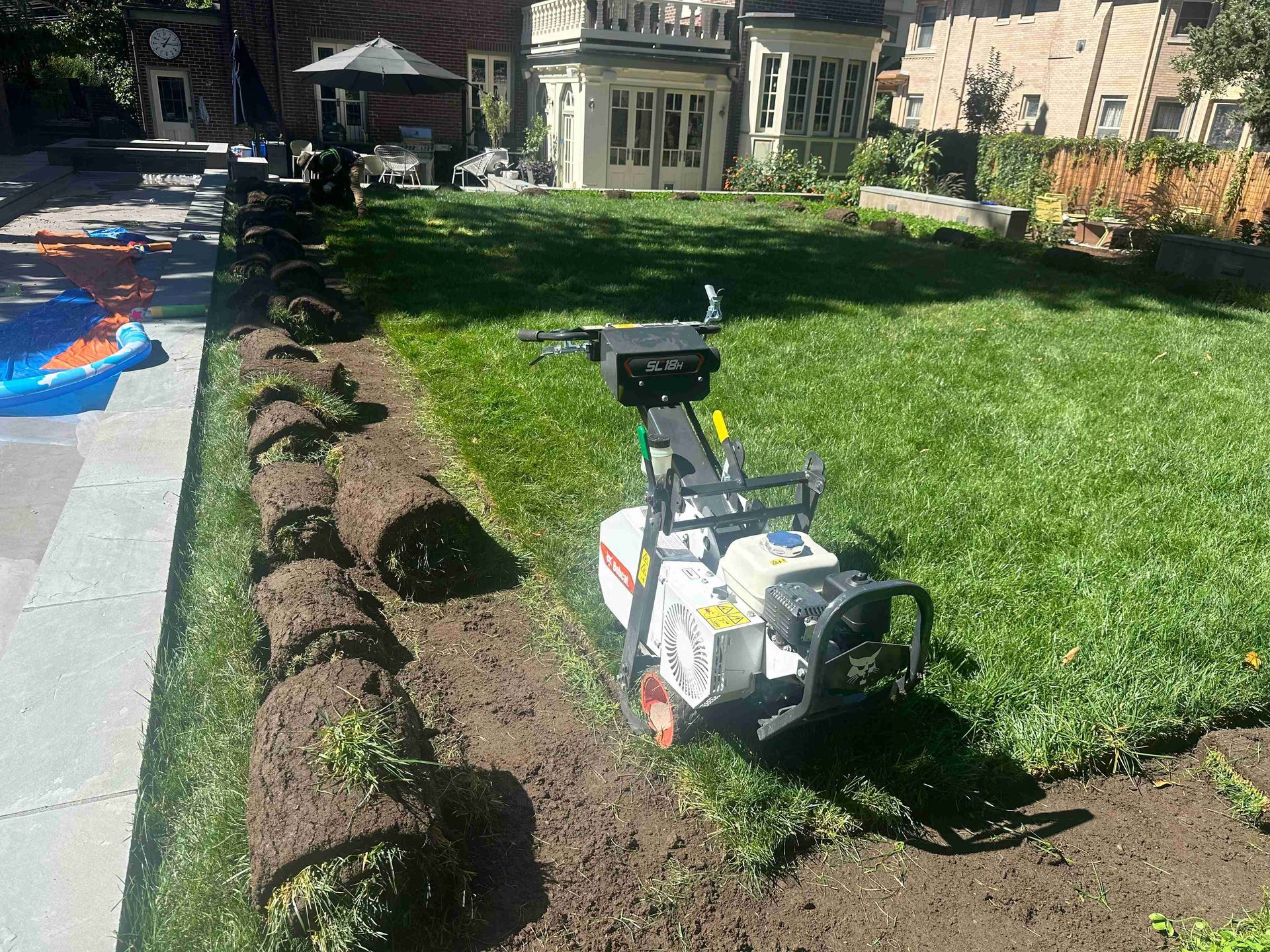 Small wheeled machine on a dirt strip beside a green lawn and trimmed shrubs in a backyard garden