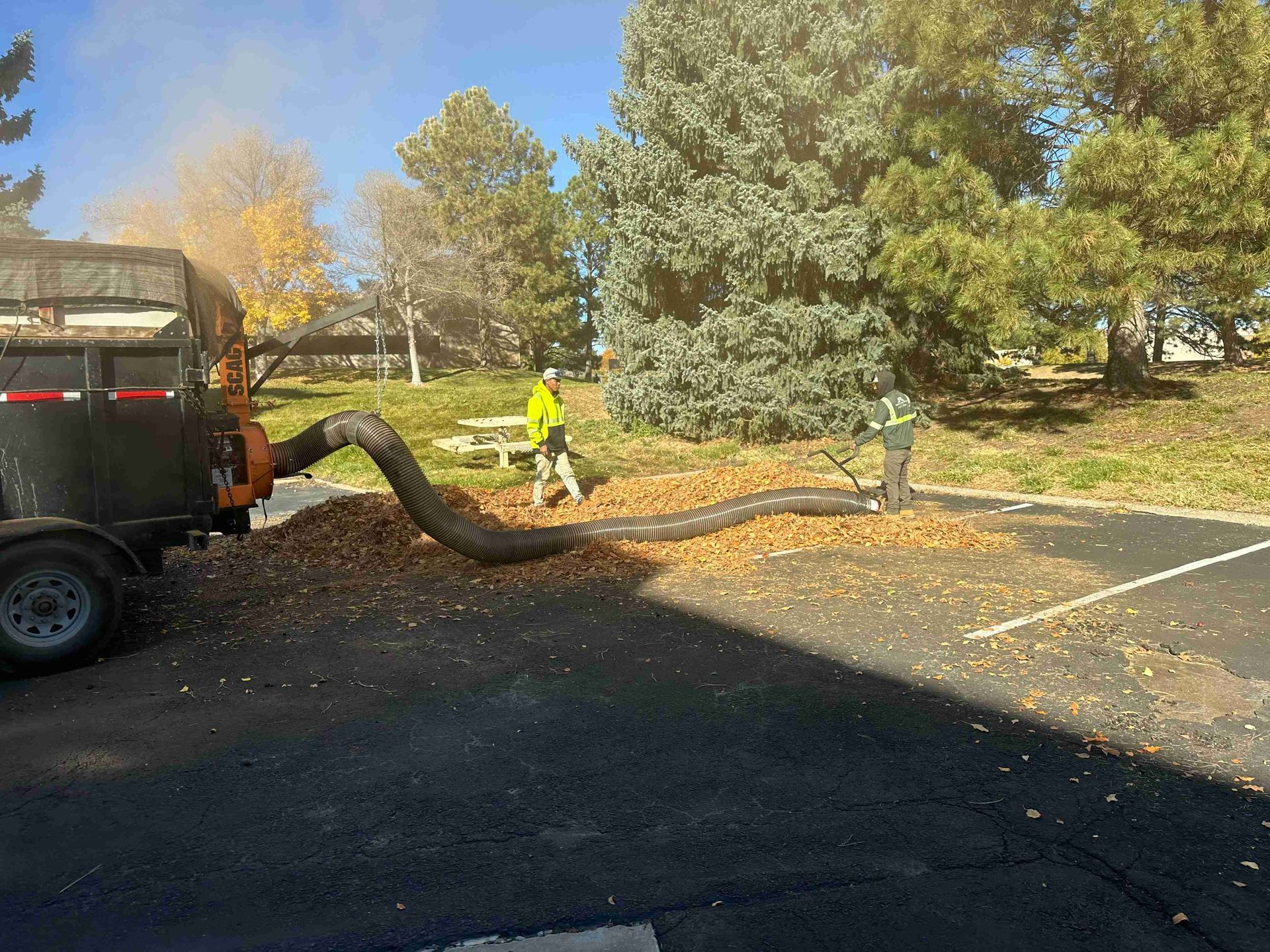 Workers using a truck hose to blow leaves across a parking lot in autumn