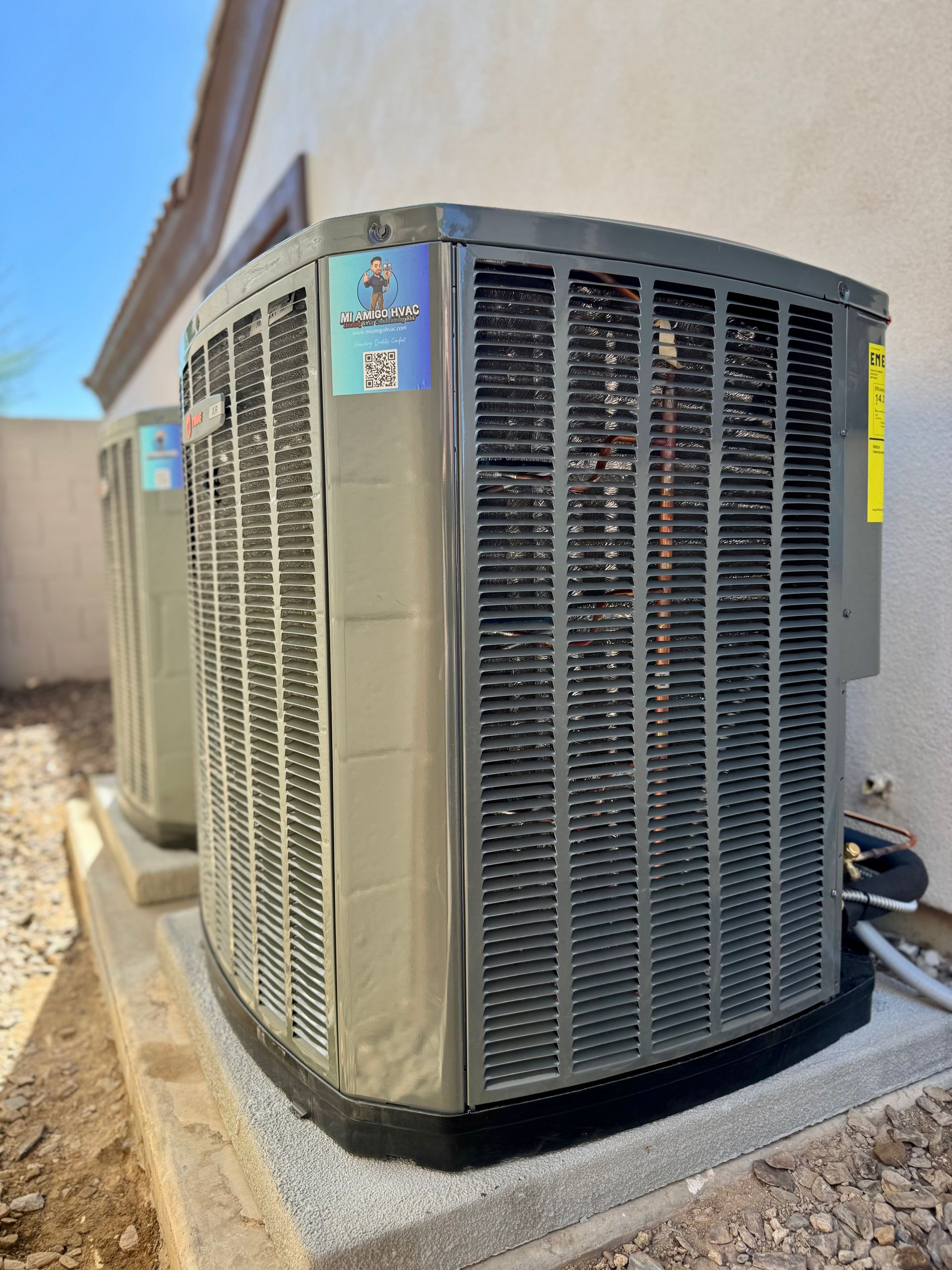 Two beige, rectangular HVAC condenser units sit on a concrete pad against a light-colored exterior wall under a blue sky.