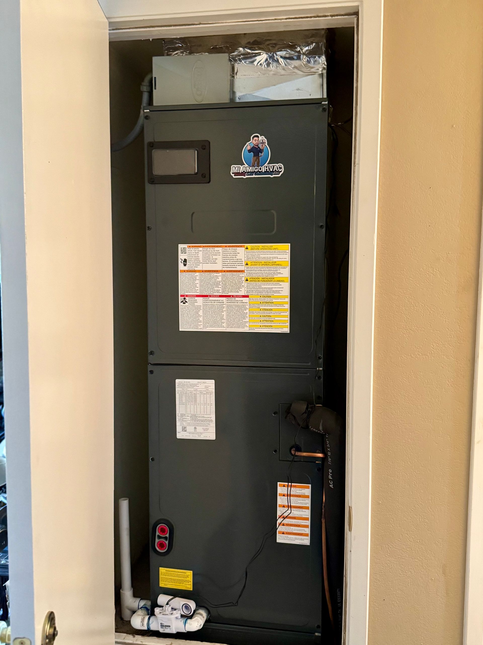 A dark gray HVAC air handler unit installed in a small, narrow closet, featuring a white condensate drain pipe at the base.