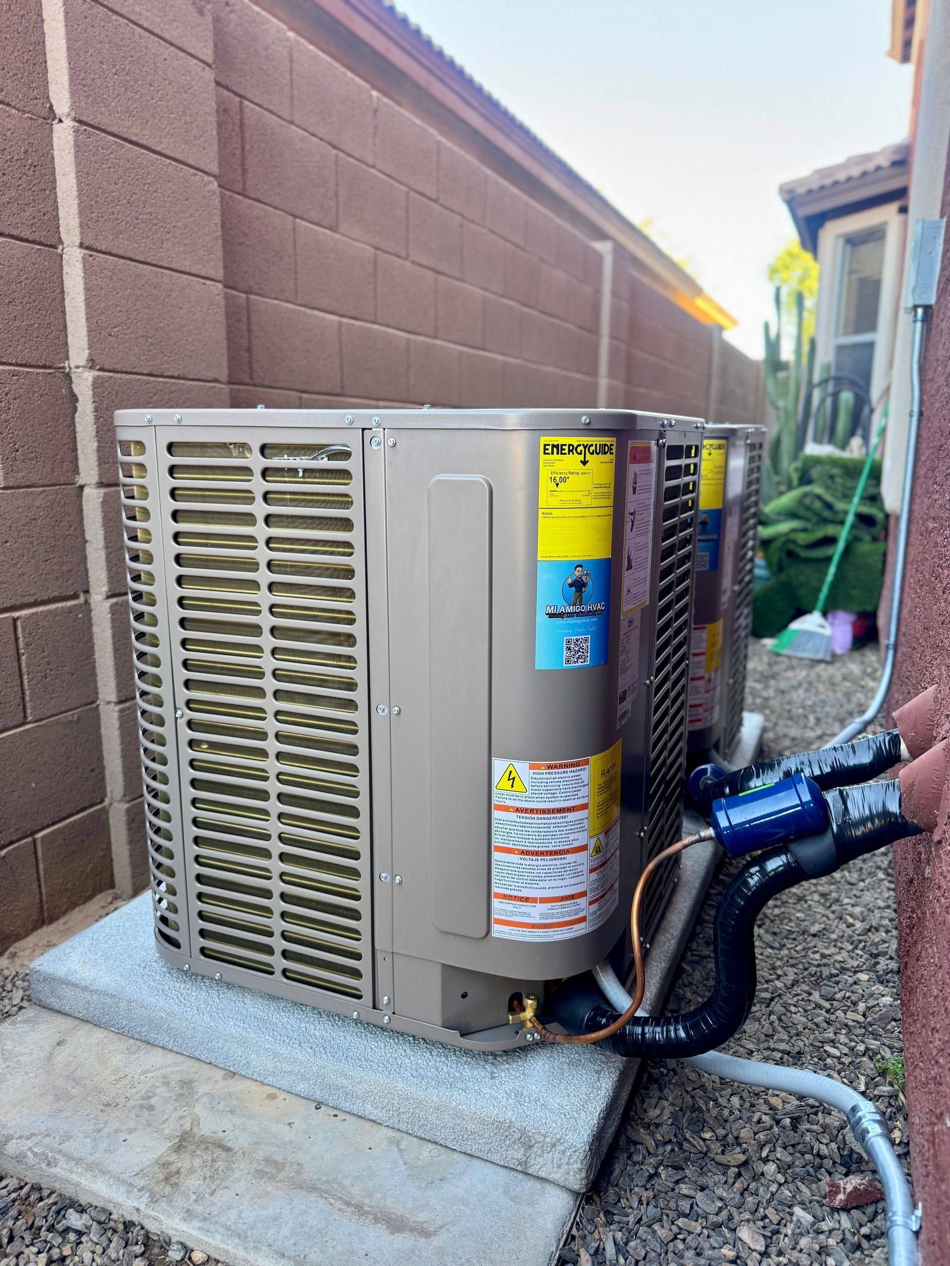 Two beige HVAC condenser units sit on a concrete slab in a narrow side yard next to a tan masonry wall.
