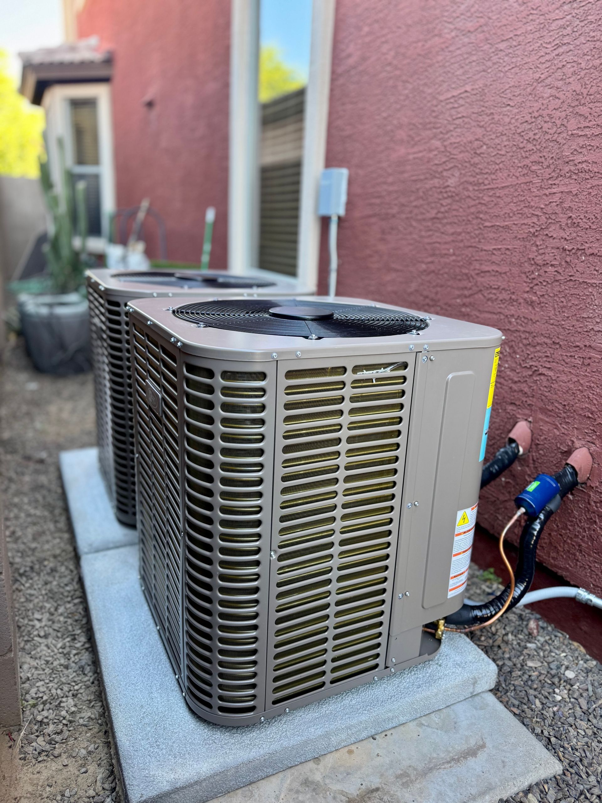 Two tan residential air conditioning units sit side-by-side on concrete pads against a reddish-brown stucco exterior wall.