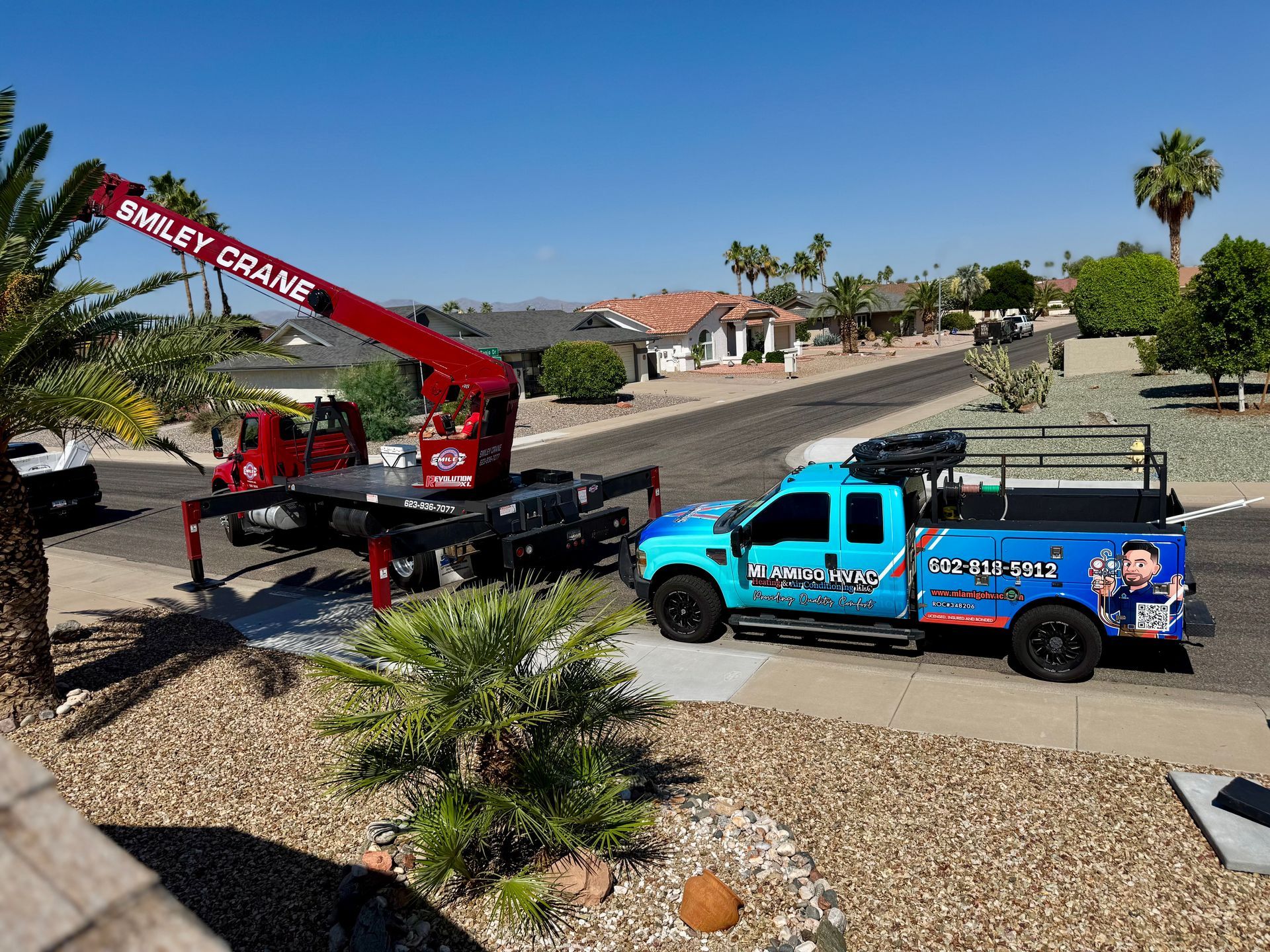 A red crane trailer and a blue service truck parked on a residential street during a sunny day.