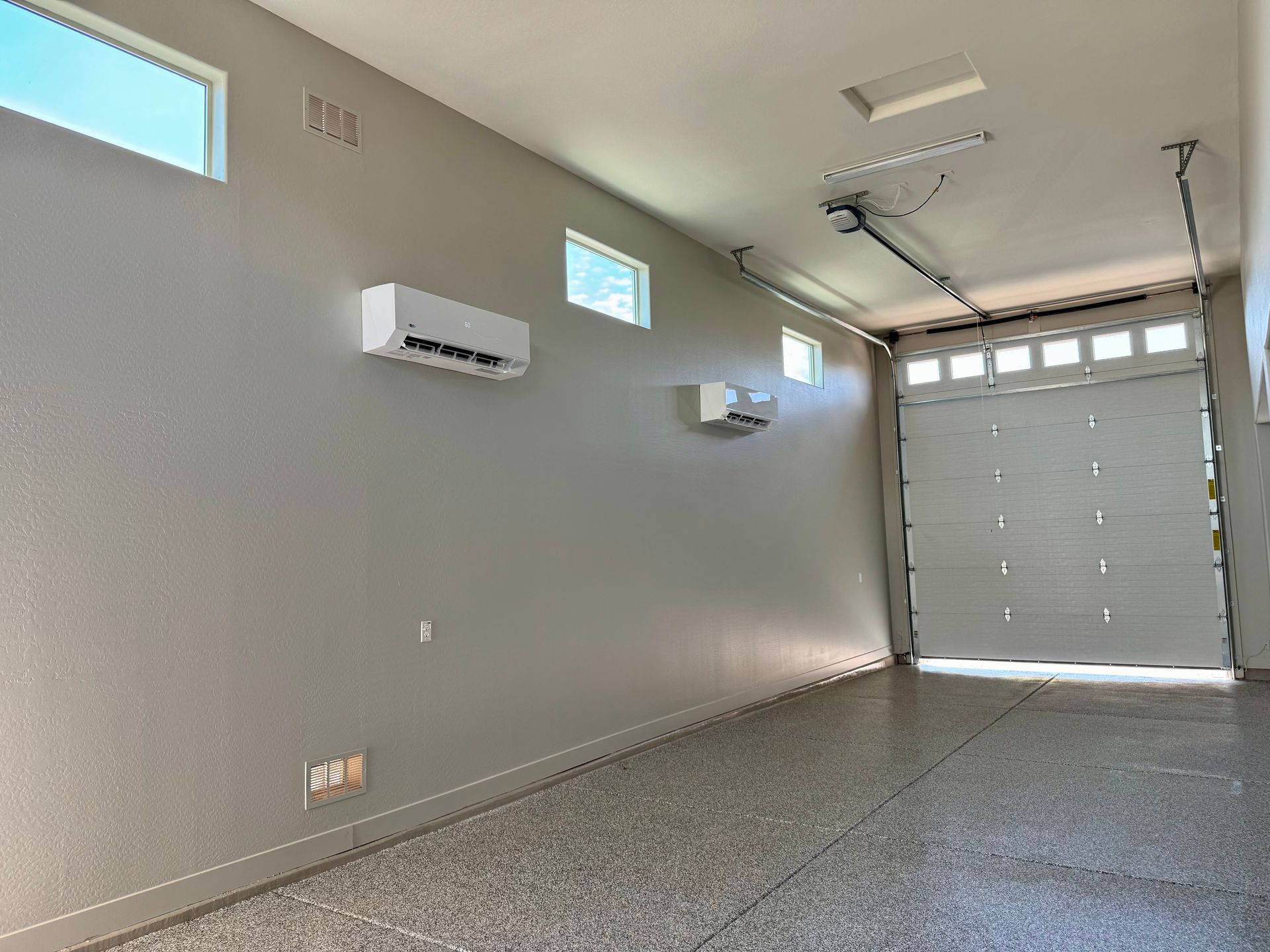 A modern garage with light gray walls, epoxy flooring, two white wall-mounted air conditioners, and a white garage door.