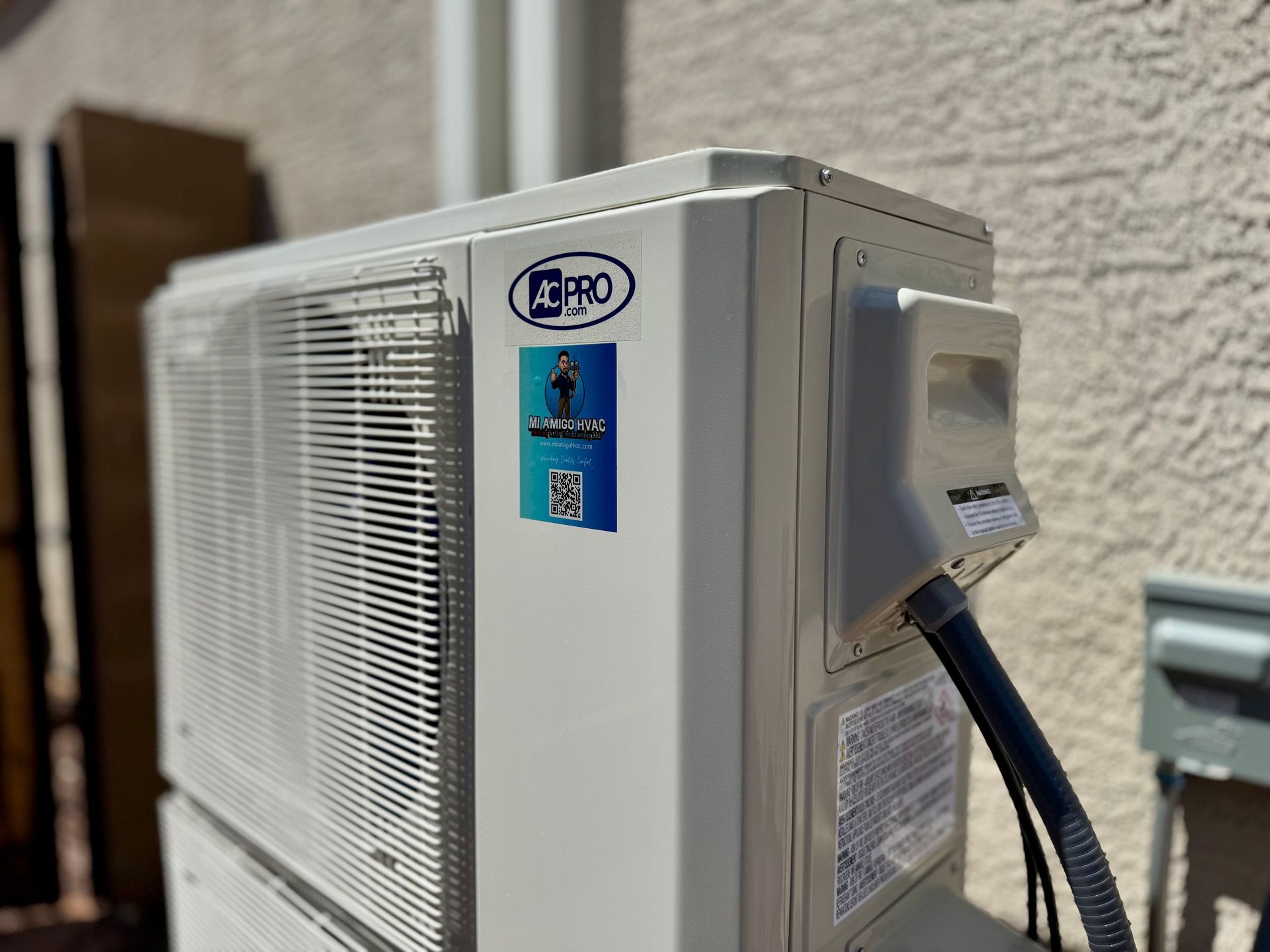 An outdoor air conditioning unit with branding stickers, connected to a power supply against a beige stucco wall.