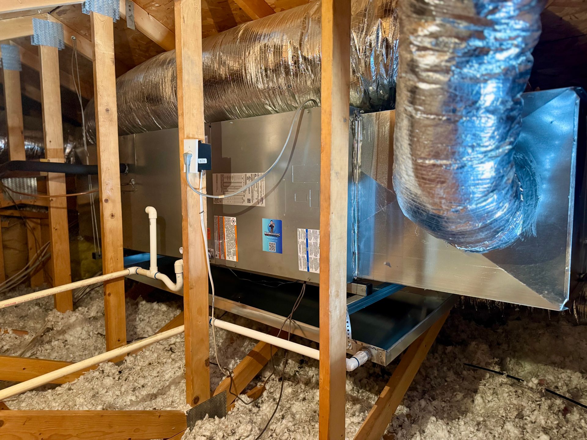 An attic HVAC air handler unit featuring silver metal casing, connected ductwork, and a white plastic condensate drain pipe.