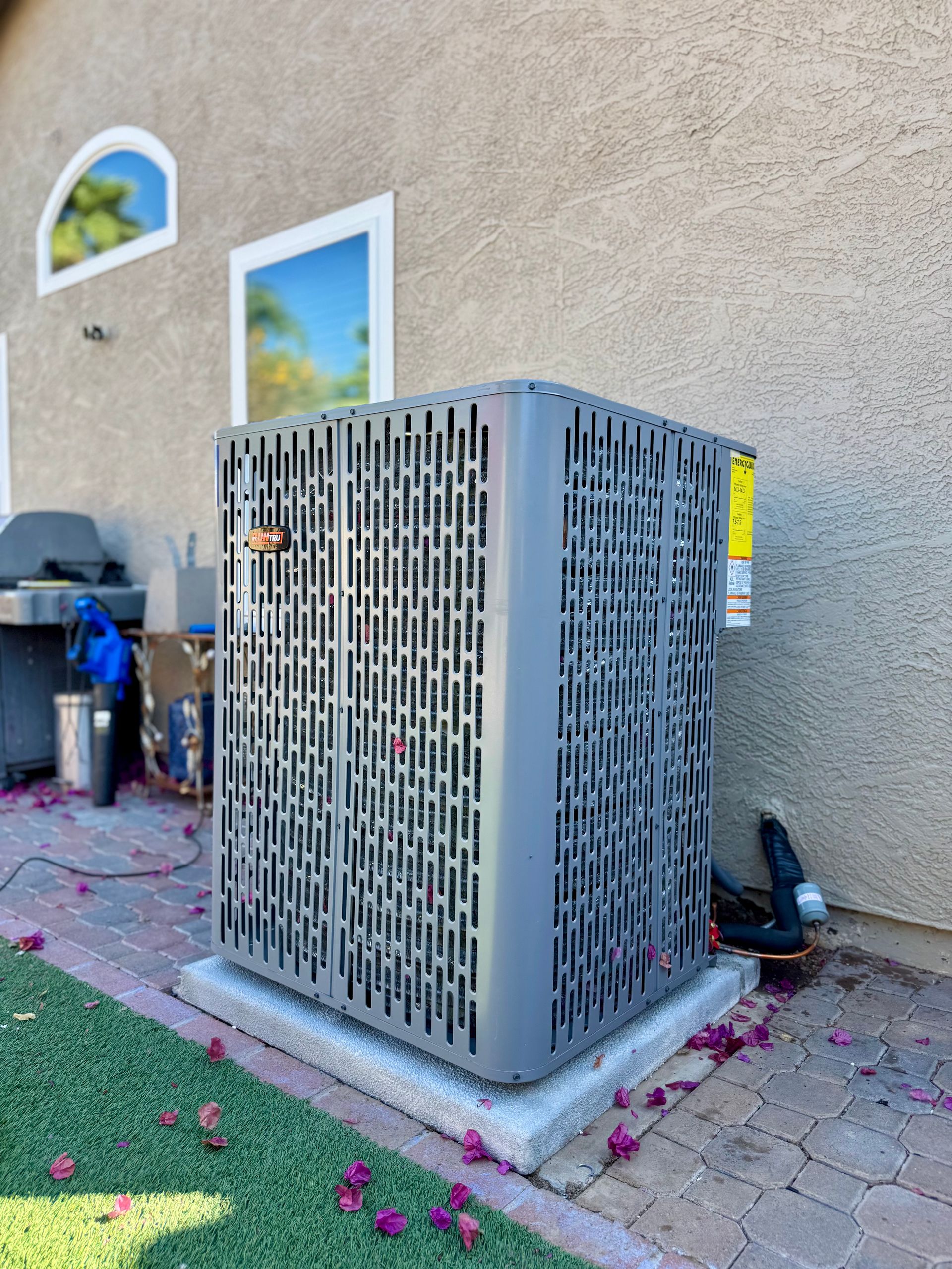 A gray residential air conditioning condenser sits on a concrete pad outside a stucco house with windows.