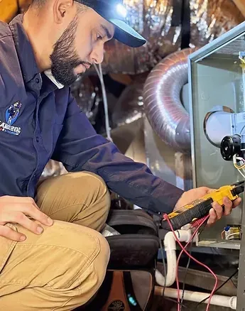 A grey residential air conditioning unit sits on a concrete slab against a brick wall next to an electrical disconnect box.