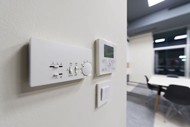 A white wall-mounted thermostat and control panel inside an office with a blurred table and chairs in the background.