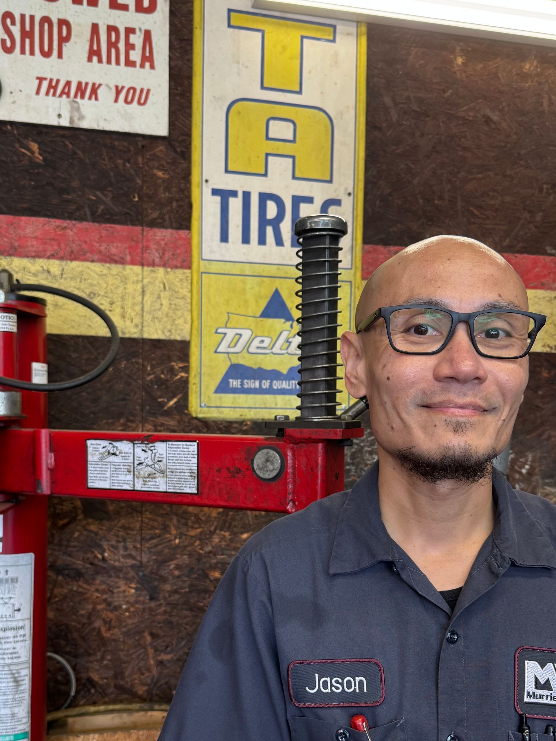 Man in glasses standing by a red tire-changing machine in a shop, with a yellow “TIRES” sign behind him