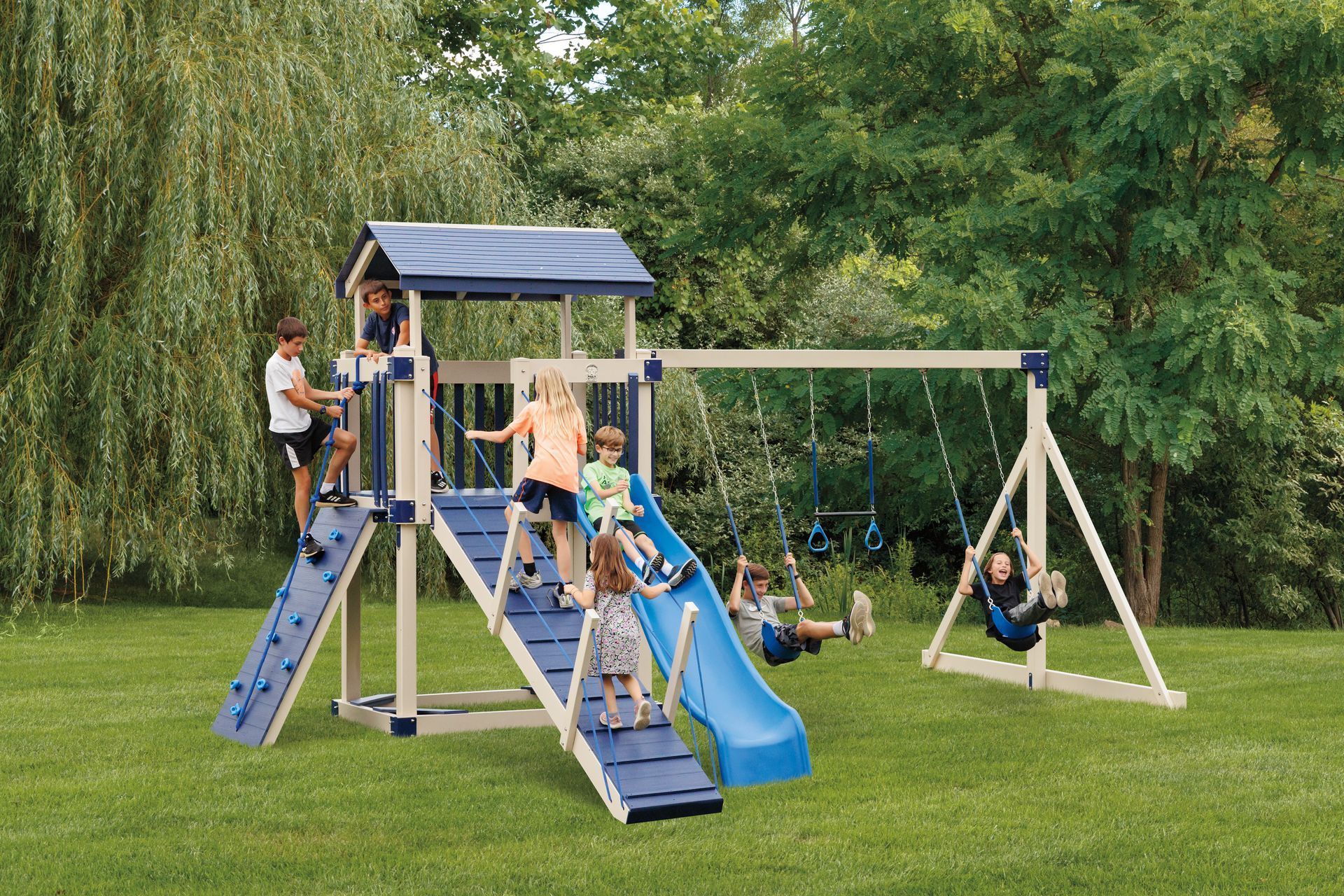 Children playing on a blue-and-beige wooden playset with a slide, climbing wall, and swings in a grassy yard.