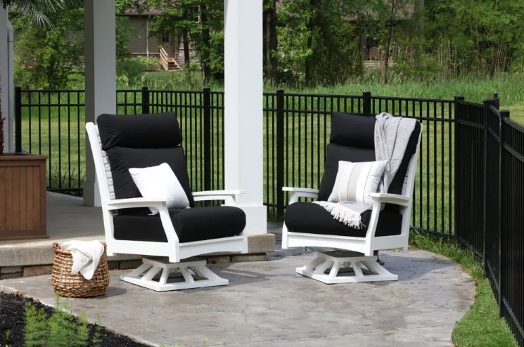 Two black and white outdoor swivel chairs on a patio, with pillows and a woven basket, near a black fence.