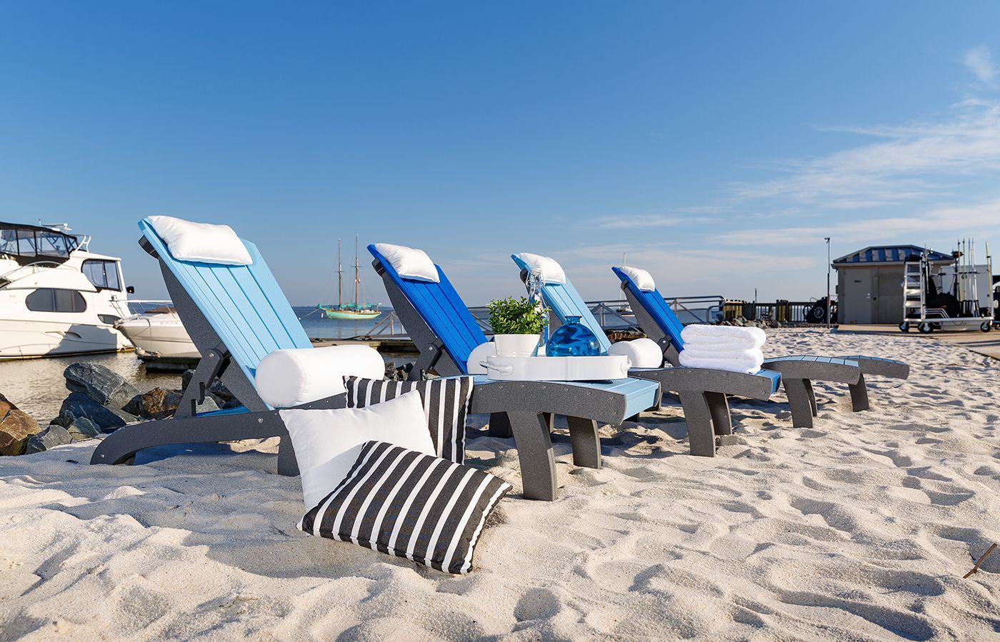 Chaise lounge chairs on sand with pillows, blue sky, and harbor background.