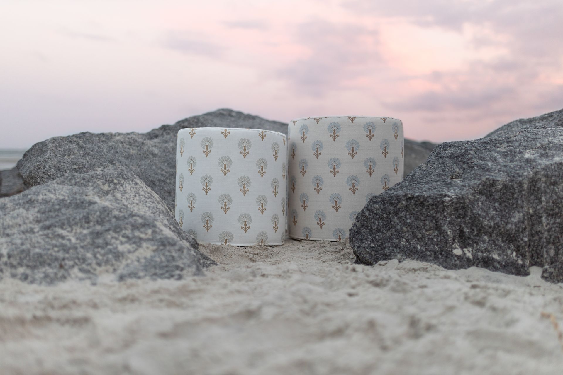 Two cushions with floral patterns sit between rocks on a beach at sunset.