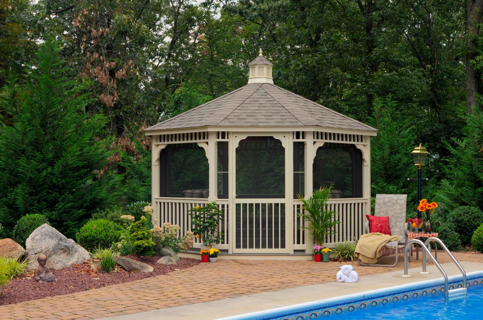 White gazebo with screened openings and brown roof near a pool surrounded by greenery.