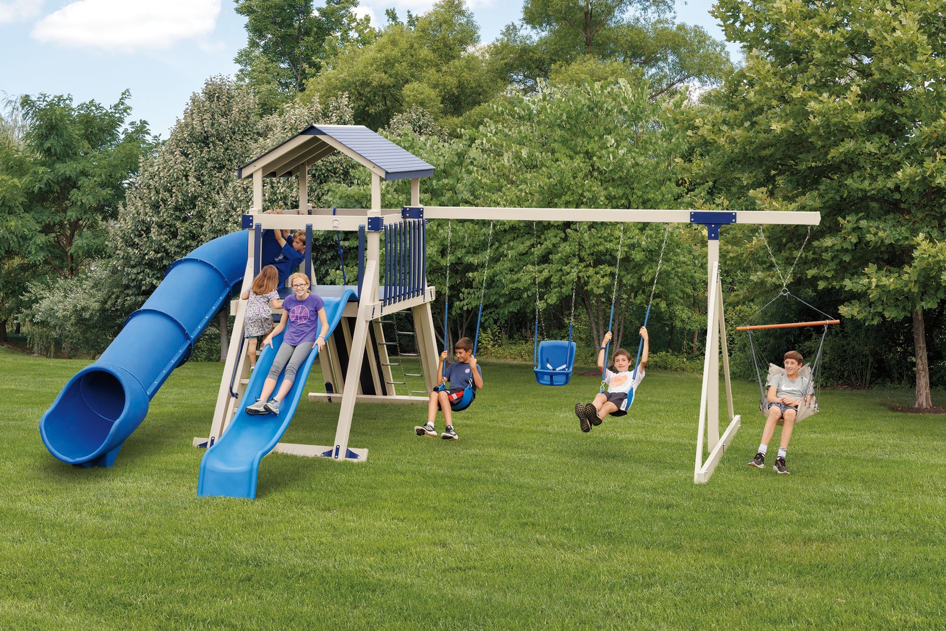 Children playing on a blue and beige wooden playground set with a slide and swings in a grassy yard.