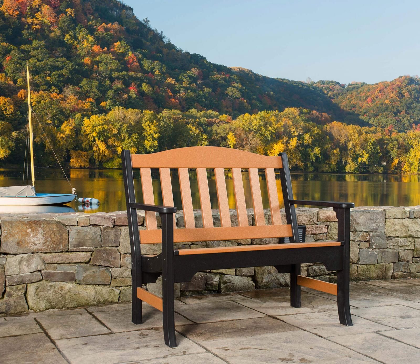 Bench with brown and black slats on a stone patio overlooking a lake with fall foliage.