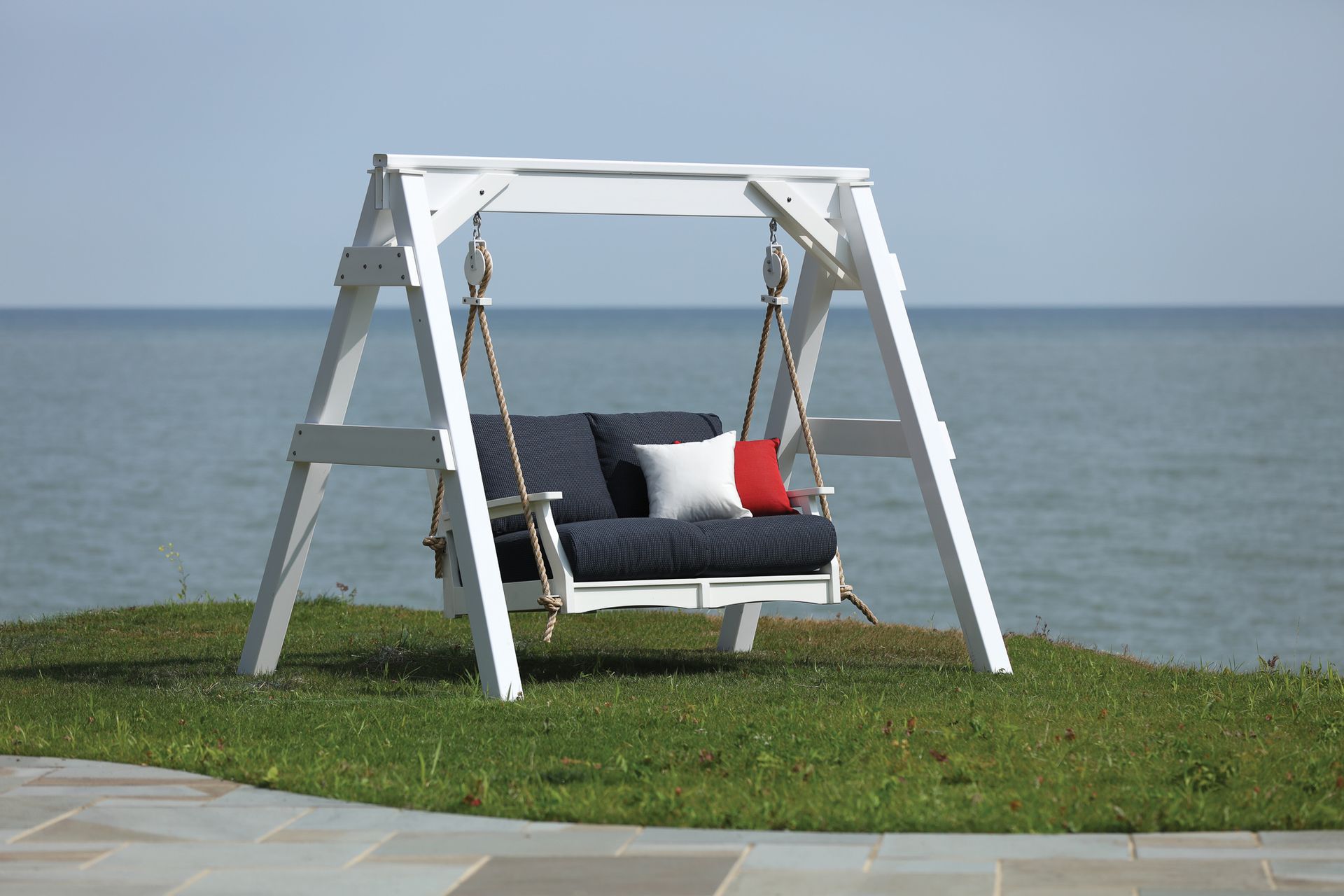 White wooden swing seat with blue cushions, red and white pillows, set on grass overlooking water.