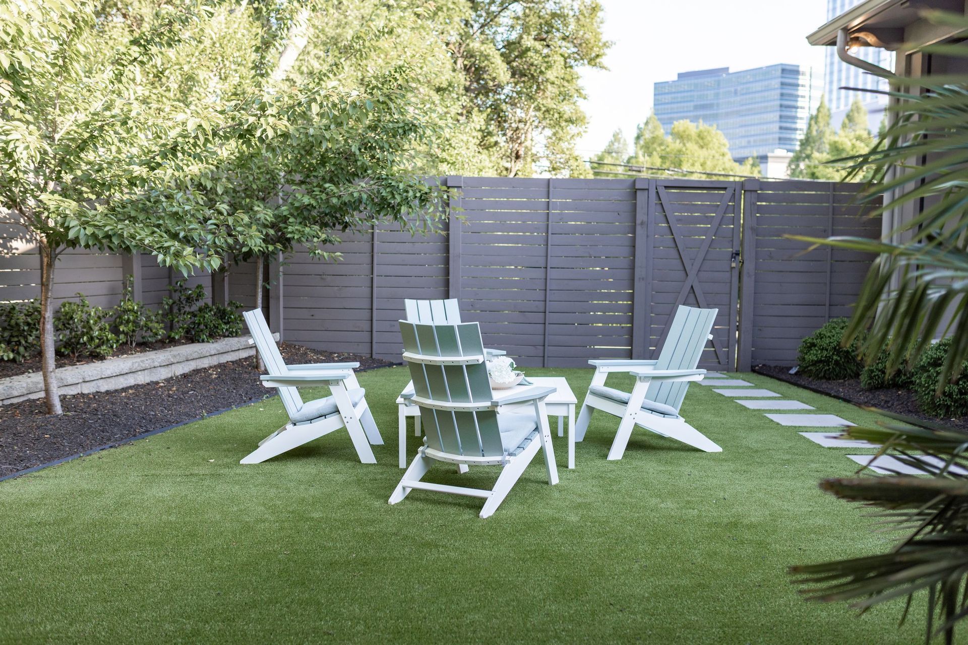 A backyard seating area with green artificial turf, gray fence, white chairs, and a small table.