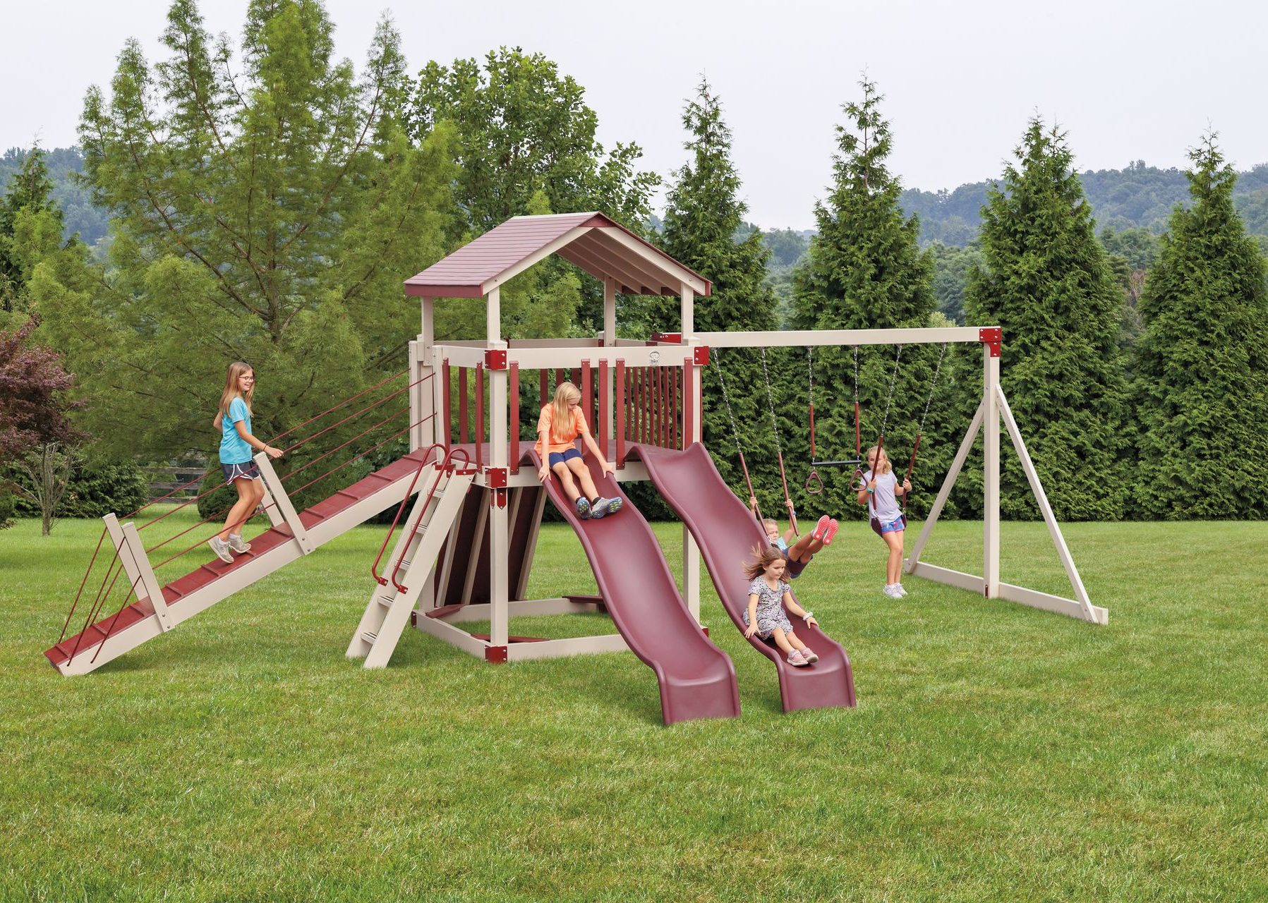 Children playing on a blue-and-beige wooden playset with a slide, climbing wall, and swings in a grassy yard.