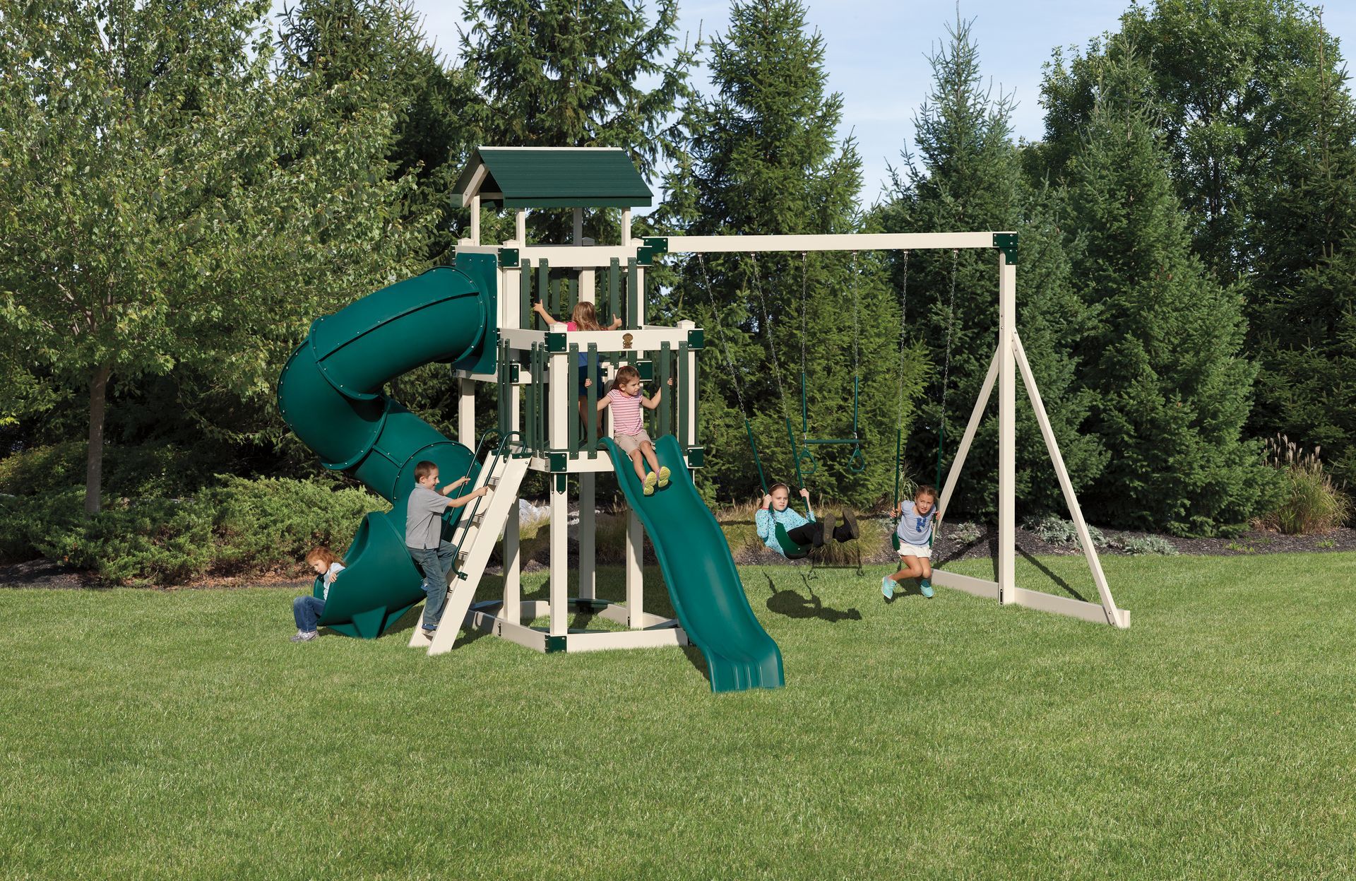 Children playing on a green and beige playset with a slide, swings, and climbing structure in a grassy yard.