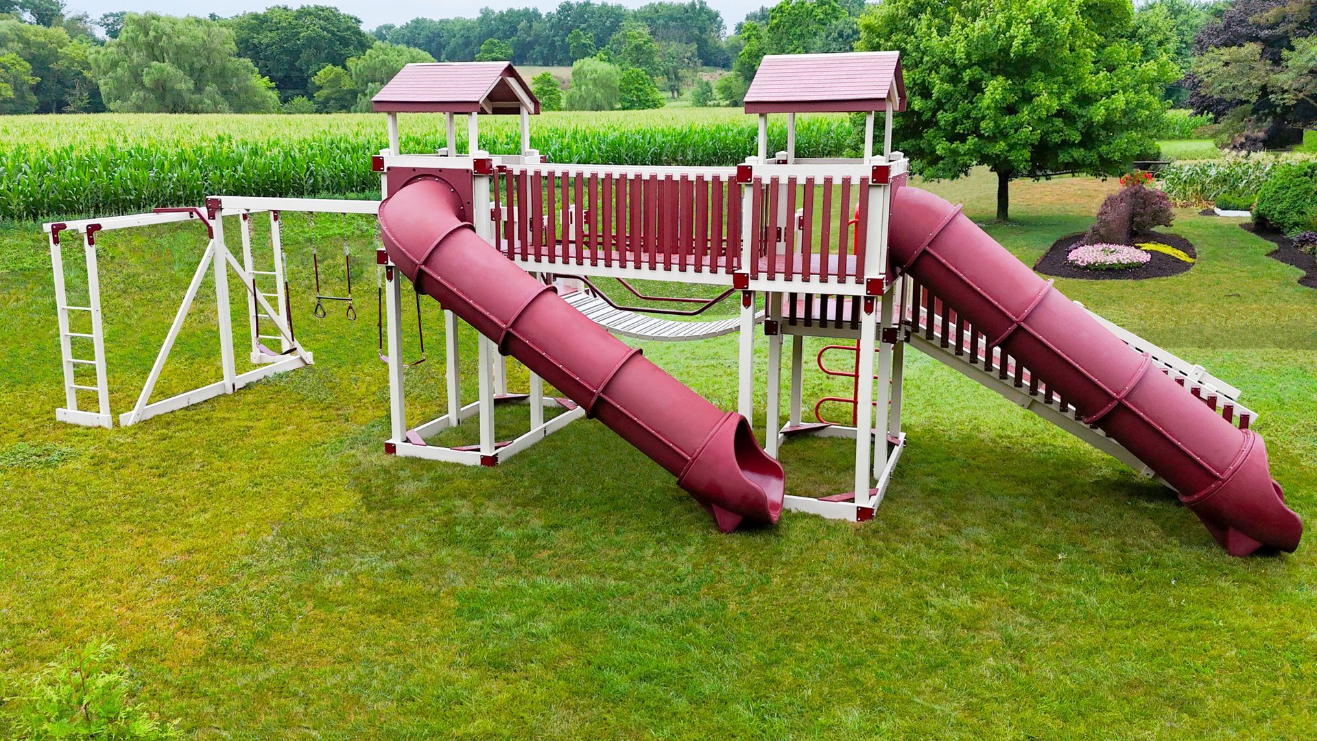 Playground set with red slides, brown roof, and white structure in a grassy yard.