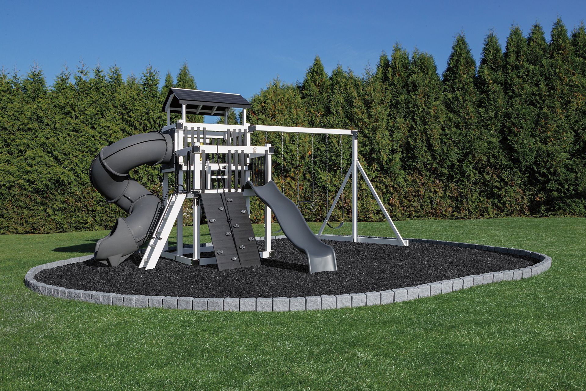 Playground structure with slide, swings, and climbing elements in a grassy yard, surrounded by black mulch.