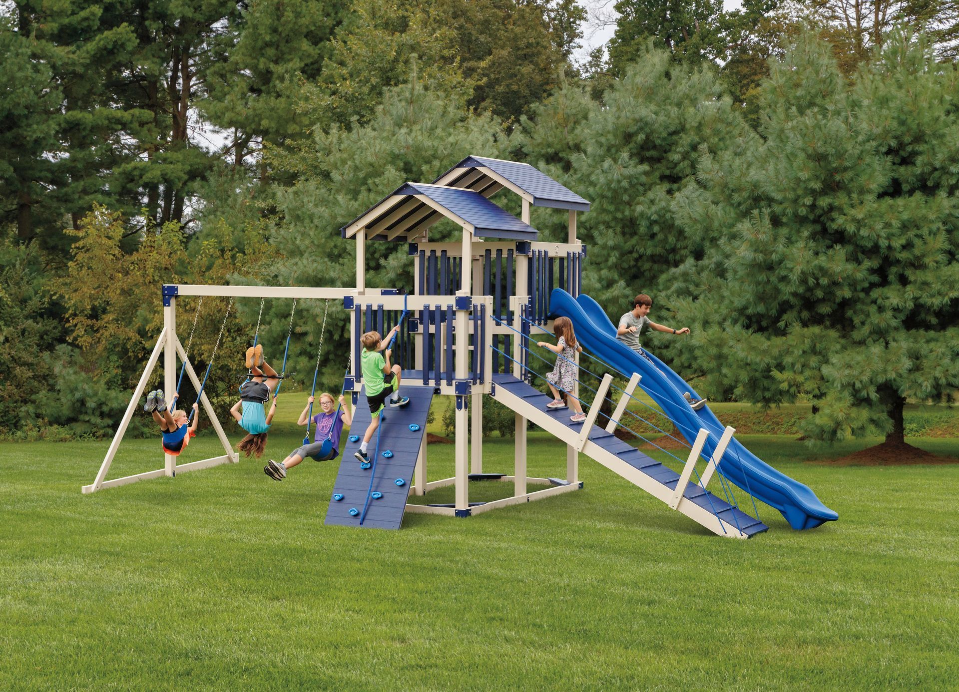 Children playing on a wooden swing set with a slide and climbing wall in a grassy yard.