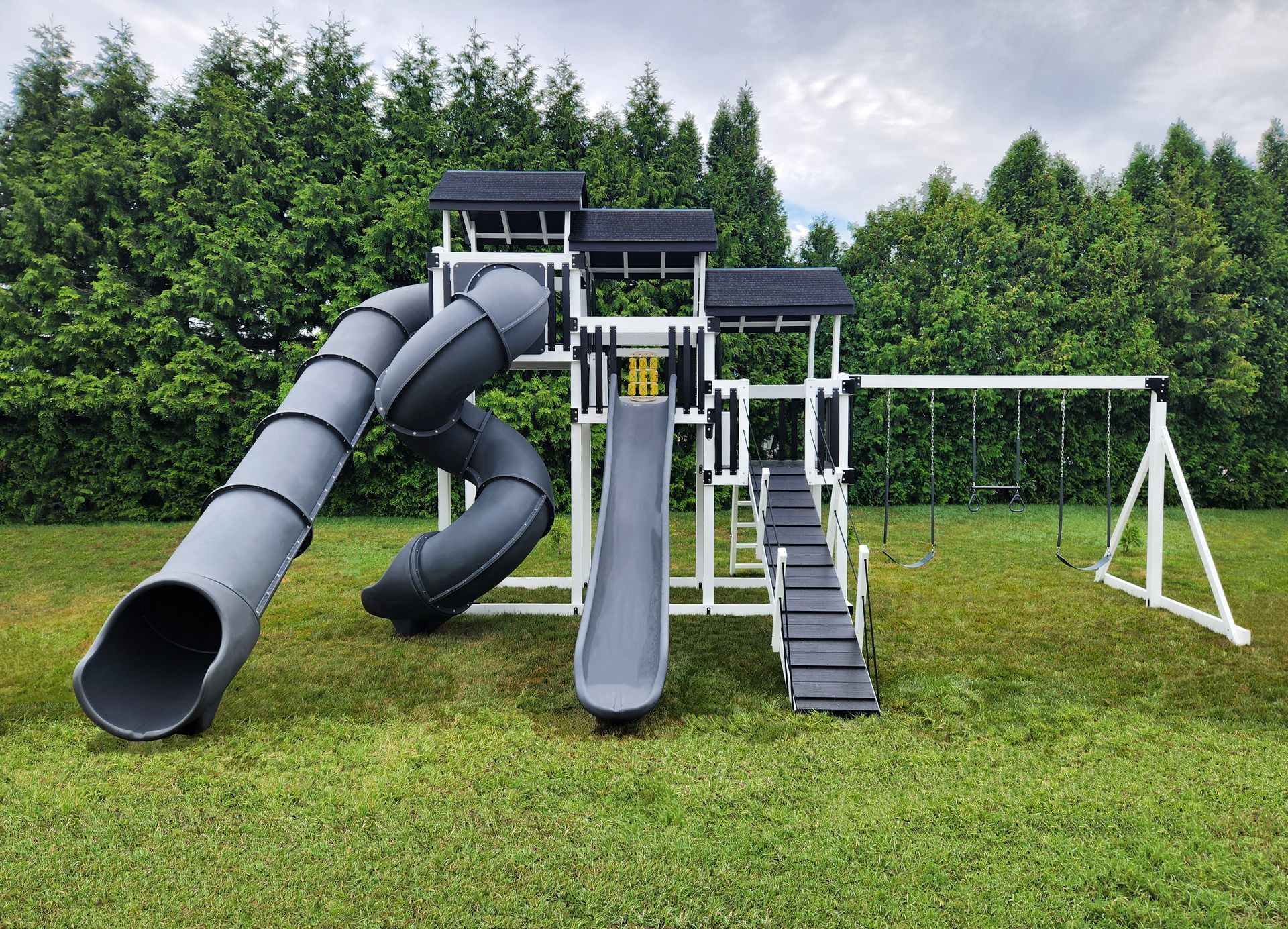 Playground with gray slides, swing set, and steps, on green grass, backed by trees.