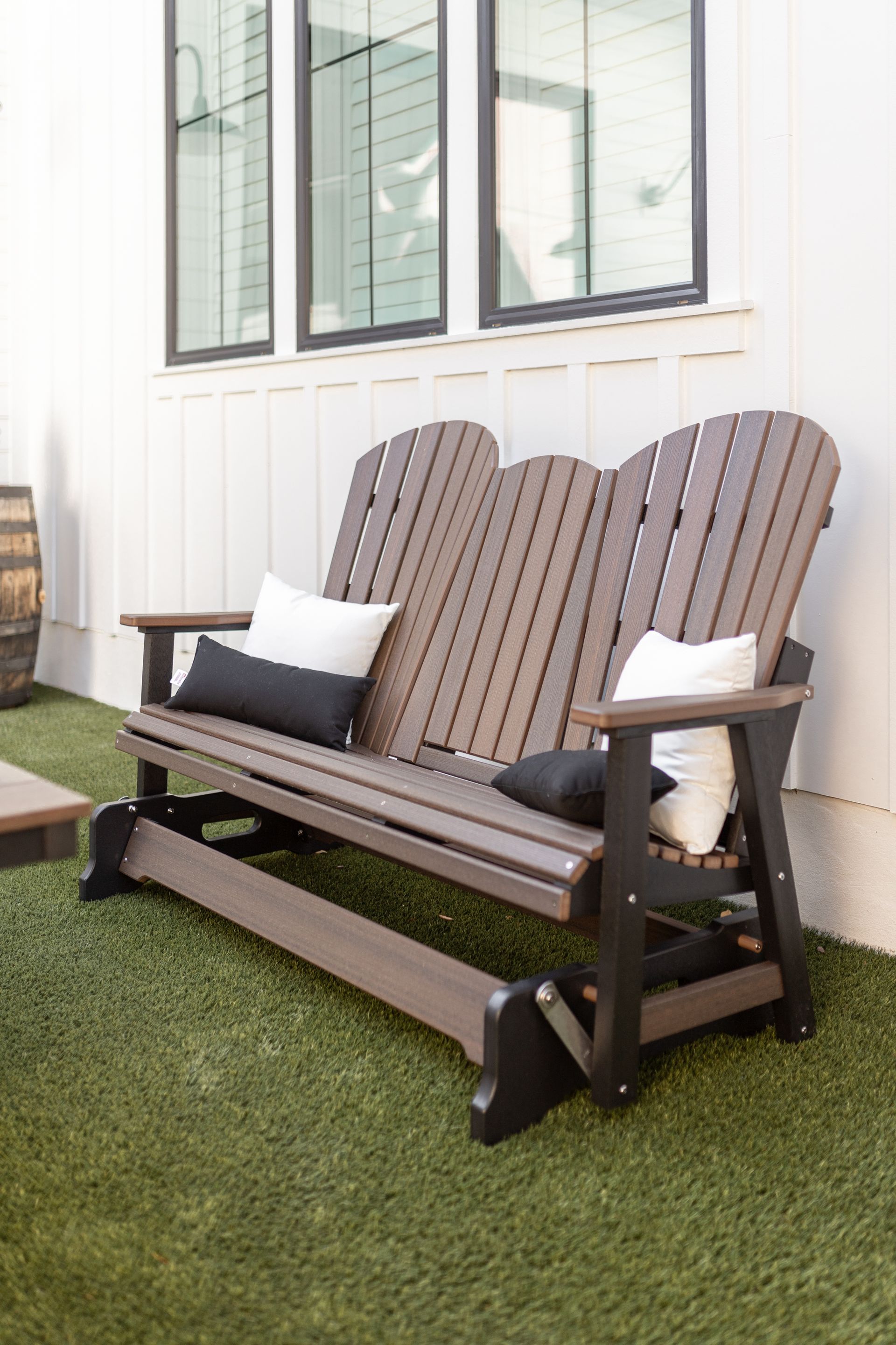 Brown and black glider bench with cushions on green turf, near a white wall and windows.