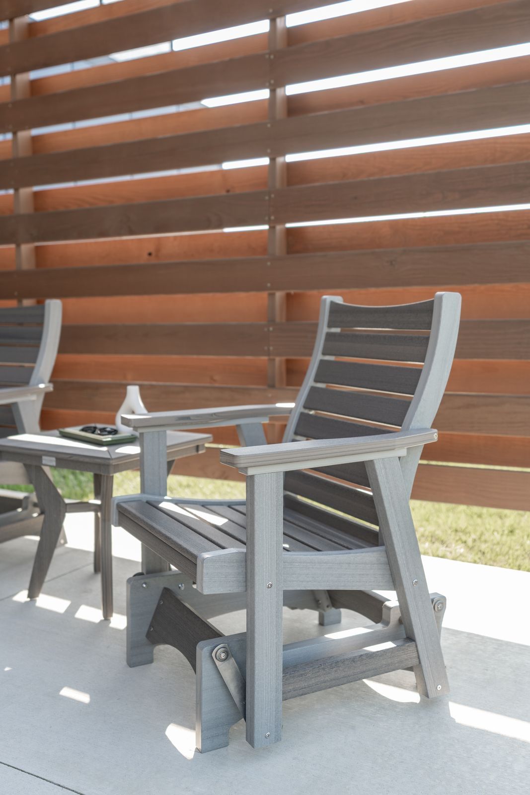 Gray outdoor glider chair and small table on concrete patio with horizontal wood fence in background.